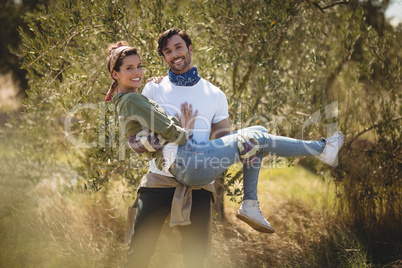 Smiling young man carrying girlfriend by trees at farm