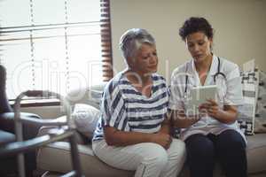Female doctor discussing over digital tablet with senior woman in living room