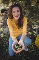 Smiling woman showing olives while crouching on field at farm