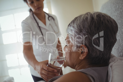 Female doctor giving sick senior woman a glass of water on bed