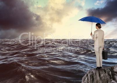 Rear view of businesswoman standing on rock holding blue umbrella and briefcase looking at sea again