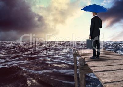 Rear view of businessman holding blue umbrella with briefcase and looking st sea while standing on p