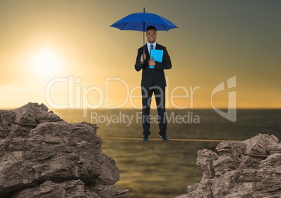 Digital composite image of businessman standing on rope holding diary with blue umbrella amidst rock