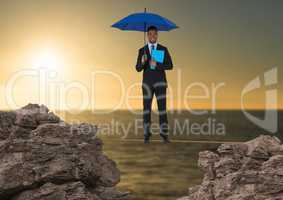 Digital composite image of businessman standing on rope holding diary with blue umbrella amidst rock