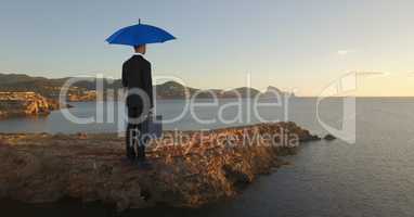 Businessman standing on rock with blue umbrella and briefcase while looking at sea