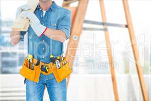 Carpenter wearing a tool belt and coverall is holding a wooden board against ladder background