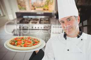 A cook wearing a chef's hat is holding a pizza on a plate against kitchen background