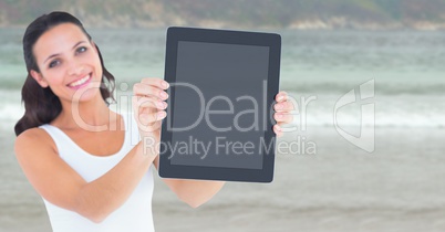 Smiling woman holding out tablet against blurry beach