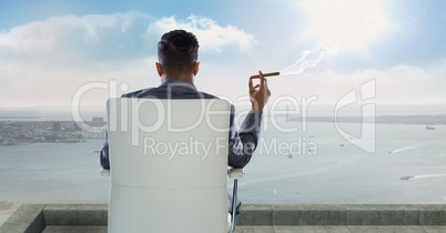Rear view of businessman sitting on chair and smoking cigar while looking at sea against sky