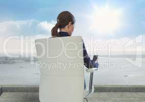 Rear view of businesswoman sitting on chair while looking at mobile phone against sky during sunny d