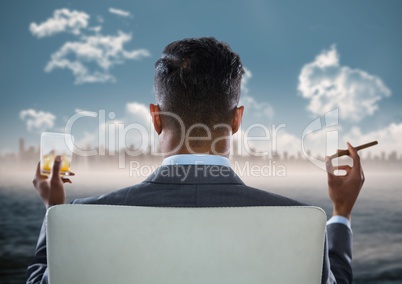 Back of seated business man smoking cigar and drinking while looking at blurry skyline and water