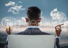 Back of seated business man smoking cigar and drinking while looking at blurry skyline and water