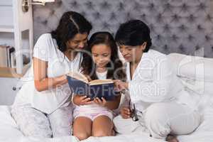 Smiling multi-generation family reading book while sitting on bed