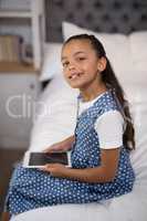 Smiling girl holding digital tablet while sitting on bed at home