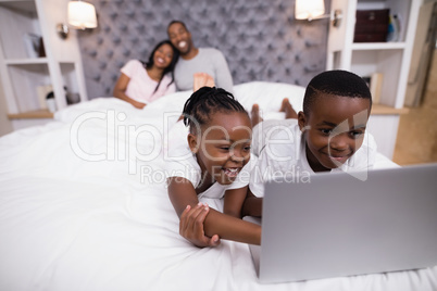 Happy siblings using laptop on bed with parents in background