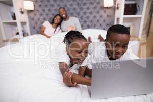 Happy siblings using laptop on bed with parents in background