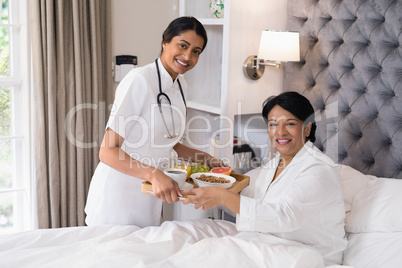 Smiling nurse serving breakfast to patient resting on bed at home