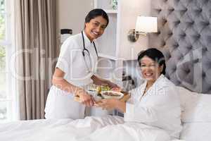 Smiling nurse serving breakfast to patient resting on bed at home