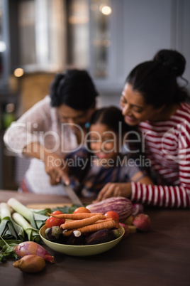 Vegetables on table against multi-generation family preparing food in kitchen