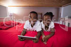 Happy siblings with digital tablet lying on bed at home