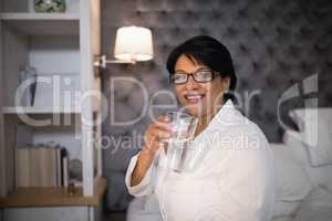 Smiling mature woman holding water glass in bedroom at home