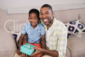 Smiling father and son with gift box sitting on sofa at home