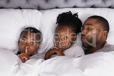 Portrait of siblings with finger on lips by father sleeping in bed