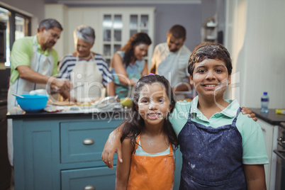 Siblings smiling at camera while family members preparing dessert in background