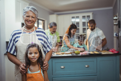Grandmother and granddaughter smiling at camera while family members preparing dessert in background