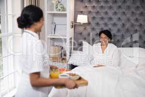 Side view of nurse serving breakfast to patient resting on bed at home