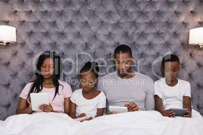 Family using digital tablets while sitting together on bed