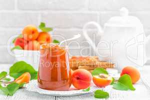 Apricot jam in a jar and fresh fruits with leaves on white wooden table, breakfast