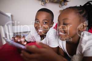 Happy siblings using mobile phone while lying on bed at home