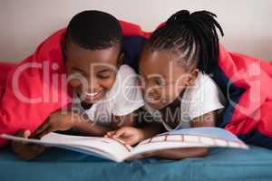 Smiling siblings reading book together while lying on bed