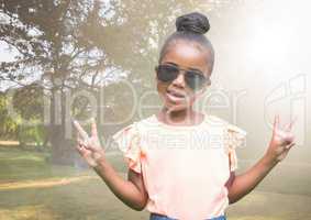 Girl in sunglasses making peace signs against blurry park with flare