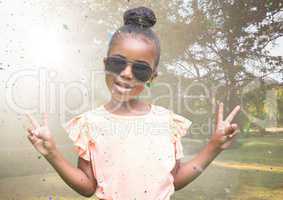 Girl in sunglasses making peace signs against blurry park with flare and confetti