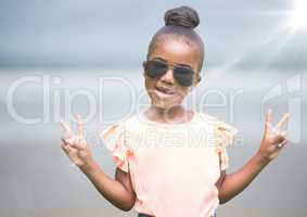 Girl in sunglasses making peace signs against blurry beach with flare