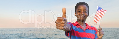 Boy with american flag giving thumbs up against water and evening sky