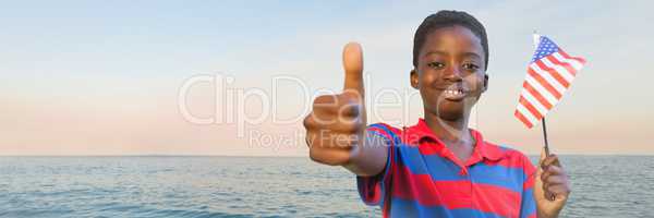 Boy with american flag giving thumbs up against water and evening sky