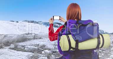 Back of millennial backpacker taking selfie against snowy hills