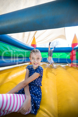 Happy girl sitting on bouncy castle while brother doing handstand in background