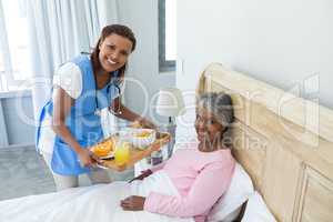 Smiling female doctor serving breakfast to senior woman on bed