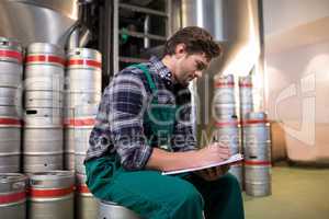 Worker writing on paper while sitting on keg at warehouse