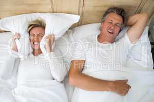 Annoyed woman covering ears with pillow while man snoring on bed