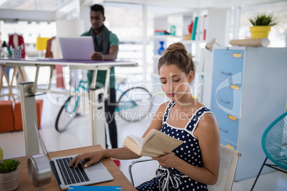 Female executive working on laptop while reading a book at desk