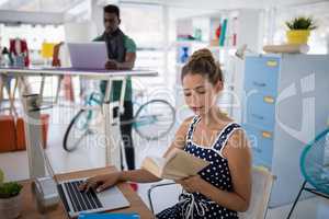Female executive working on laptop while reading a book at desk