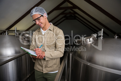 Worker amidst storage tanks writing on clipboard at brewery