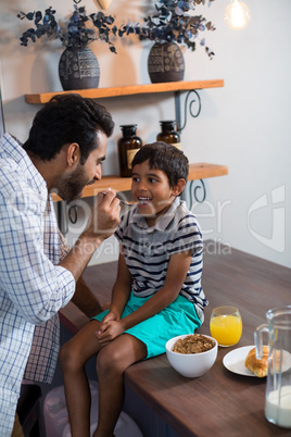 Father feeding cereal breakfast to son sitting on table