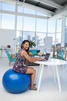 Female executive using laptop while sitting on exercise ball at desk