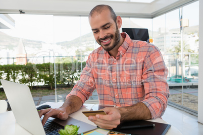 Smiling designer holding credit card while using laptop at desk